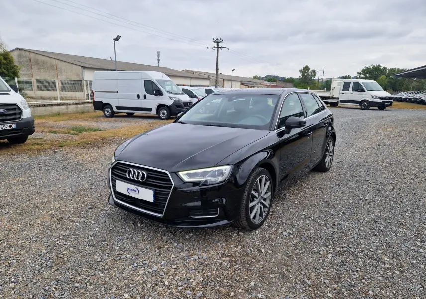 Audi A3 Sportback noir vue 3/4 avant droit sur parking gravillonné avec plusieurs véhicules utilitaires blancs en arrière-plan.