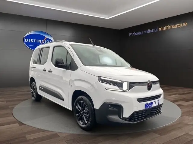 Vue 3/4 avant d'un Citroën Berlingo blanc 2025 avec jantes noires, dans un showroom au sol bois et fond gris.