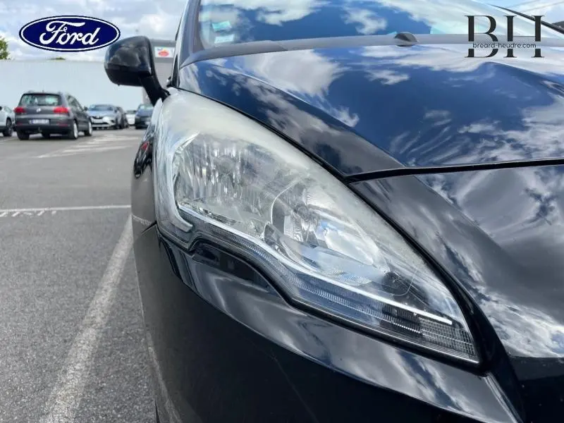 Gros plan sur le phare avant gauche d'une Peugeot 5008 noir Perla Nera, reflétant le ciel nuageux.