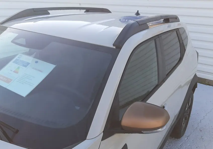 Vue 3/4 avant droit du Dacia Duster 2025 hybride en couleur Sandstone avec rétroviseur et barres de toit noirs.