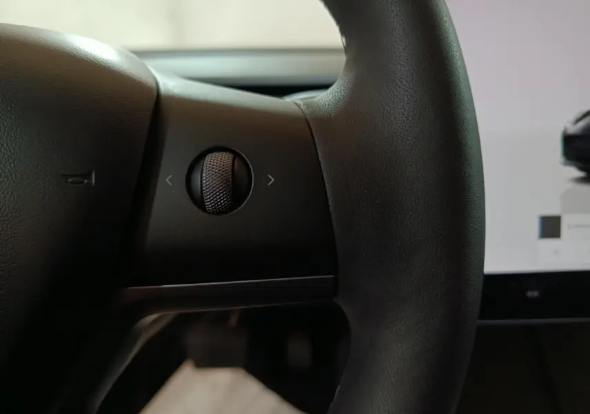 Gros plan sur la molette de commande à gauche du volant noir dans l’habitacle d’une Tesla Model 3 gris foncé.