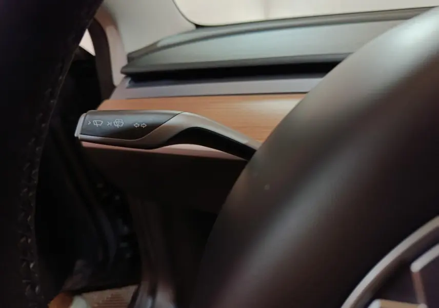 Détail du commodo d’essuie-glace sur le côté gauche du volant cuir dans l’habitacle bois clair d’une Tesla Model 3 gris foncé.