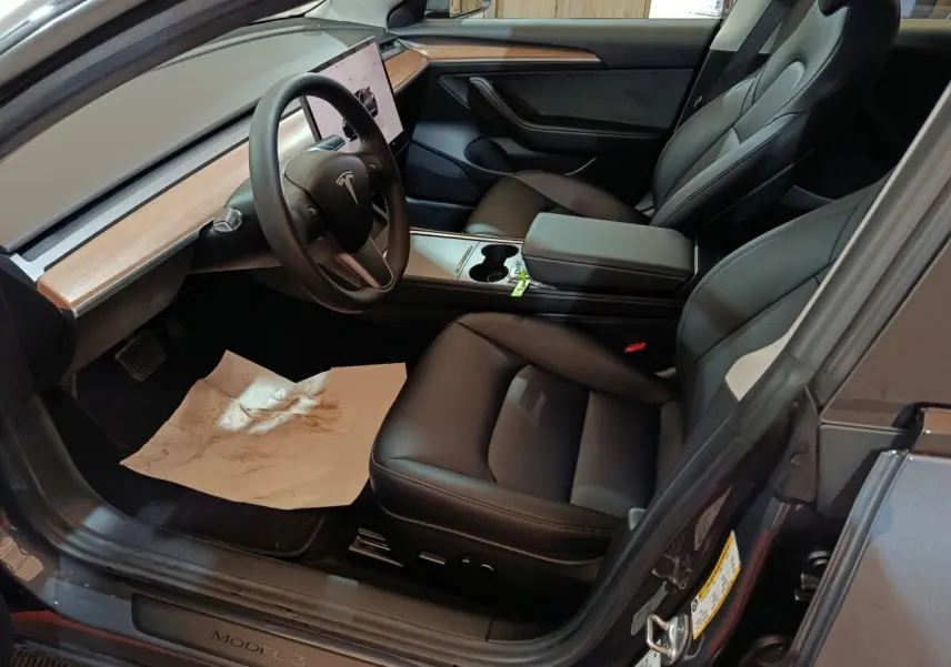 Intérieur Tesla Model 3 gris foncé, vue côté conducteur, sièges cuir noir et tableau de bord bois clair.