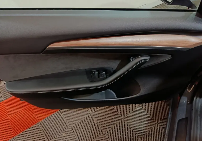 Vue intérieure côté gauche de la porte d’une Tesla Model 3 gris foncé avec garniture bois et commandes vitres.