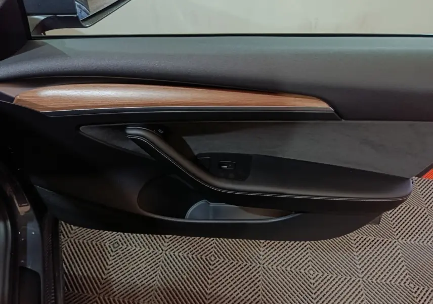 Vue intérieure du panneau de porte côté gauche de la Tesla Model 3 gris foncé, avec insert bois et poignée noire.