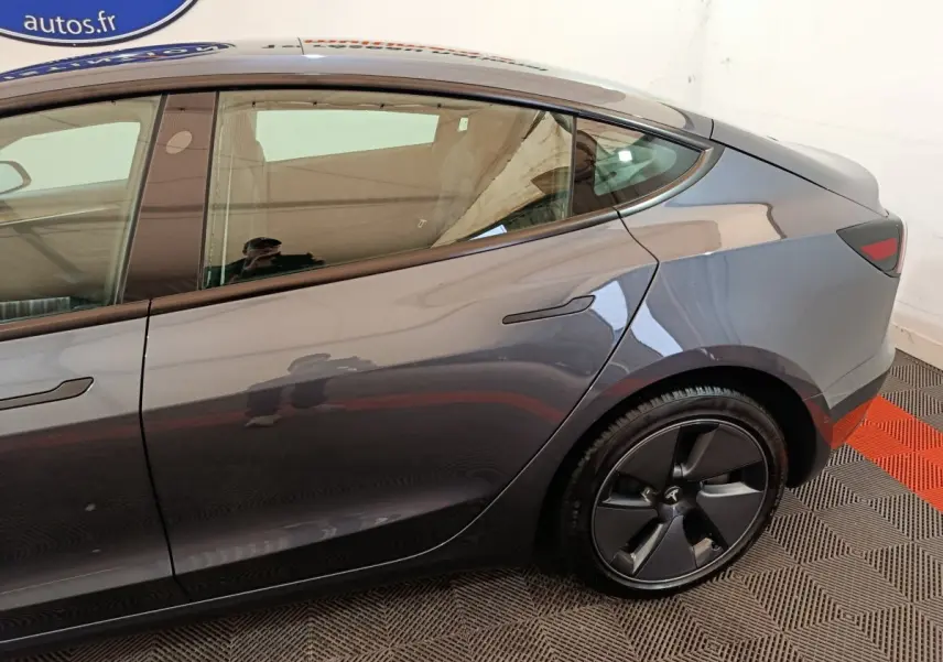 Vue latérale droite sur l'arrière d'une Tesla Model 3 gris foncé avec jante noire et poignées affleurantes.