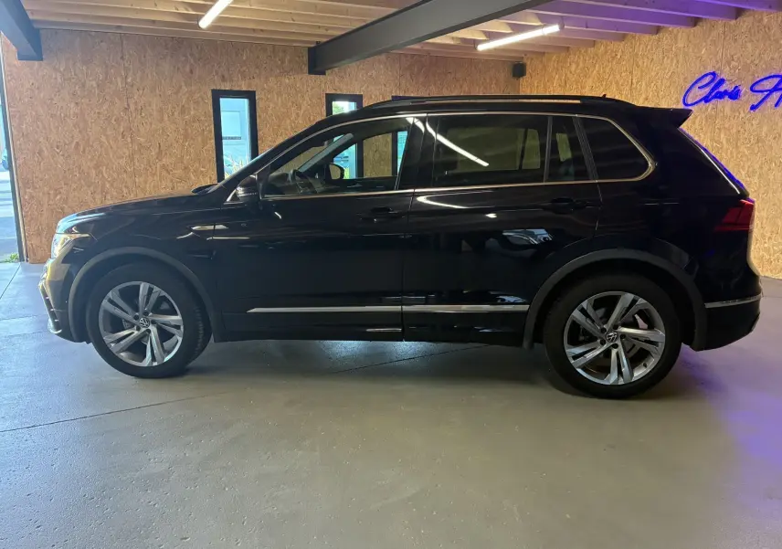 Profil côté gauche d'un Volkswagen Tiguan noir 2022 R-Line avec jantes alu, barres de toit et becquet en intérieur lumineux.