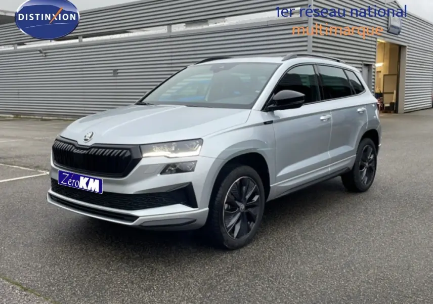 Vue 3/4 avant d'un Skoda Karoq gris argent métallisé avec jantes noires et calandre noire, stationné devant un bâtiment industriel.