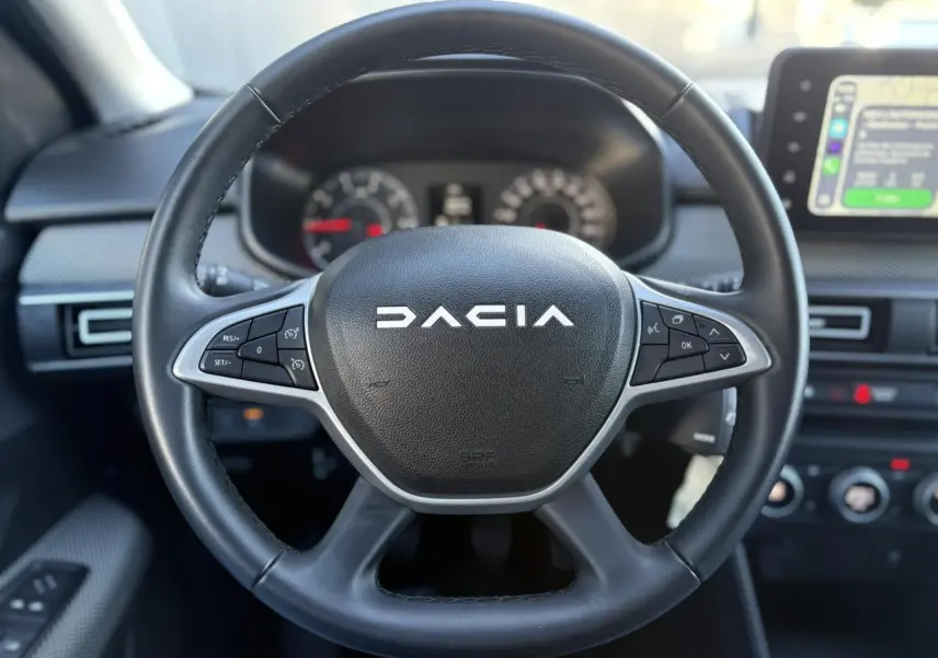Vue rapprochée du volant noir DACIA avec commandes intégrées, tableau de bord et écran tactile en arrière-plan.