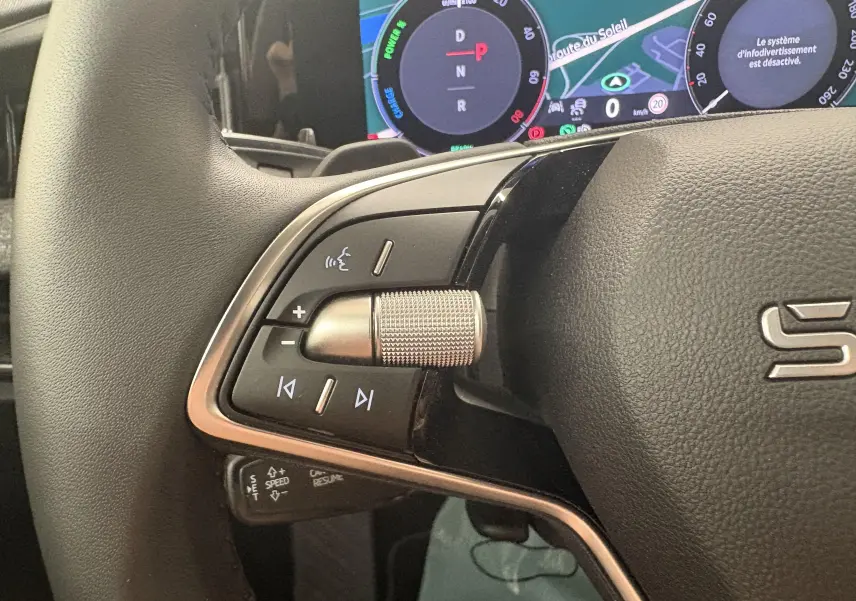 Gros plan sur les commandes gauche du volant d’un Skoda Superb Combi gris argent avec écran numérique en arrière-plan.