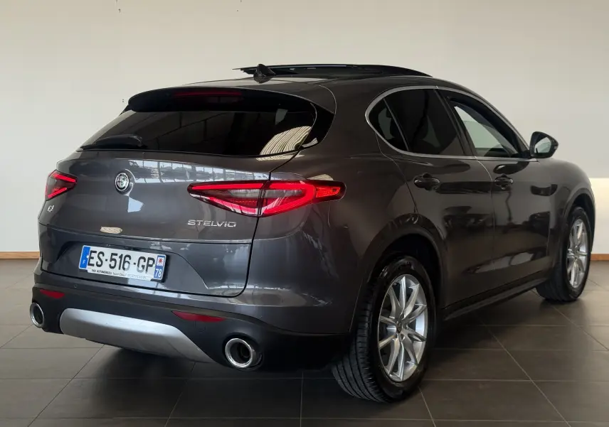 Vue 3/4 arrière droite d'un Alfa Romeo Stelvio gris 2017 avec feux arrière allumés et double sortie d'échappement.