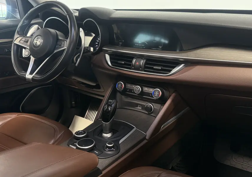 Intérieur de l'Alfa Romeo Stelvio 2017 vu côté conducteur, avec sellerie marron et tableau de bord moderne gris et bois.