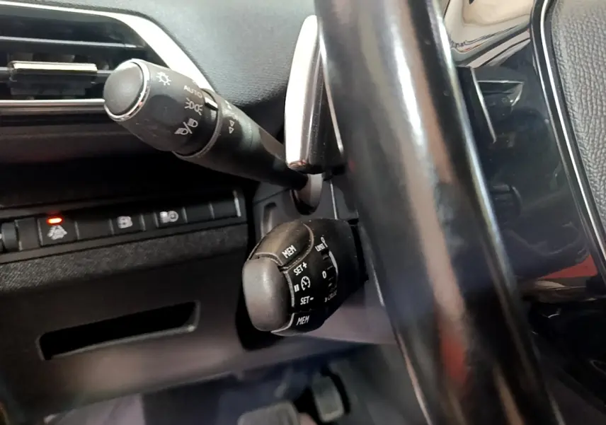 Gros plan sur la commande de régulateur de vitesse derrière le volant noir du Peugeot 3008 hybride blanc, côté gauche.