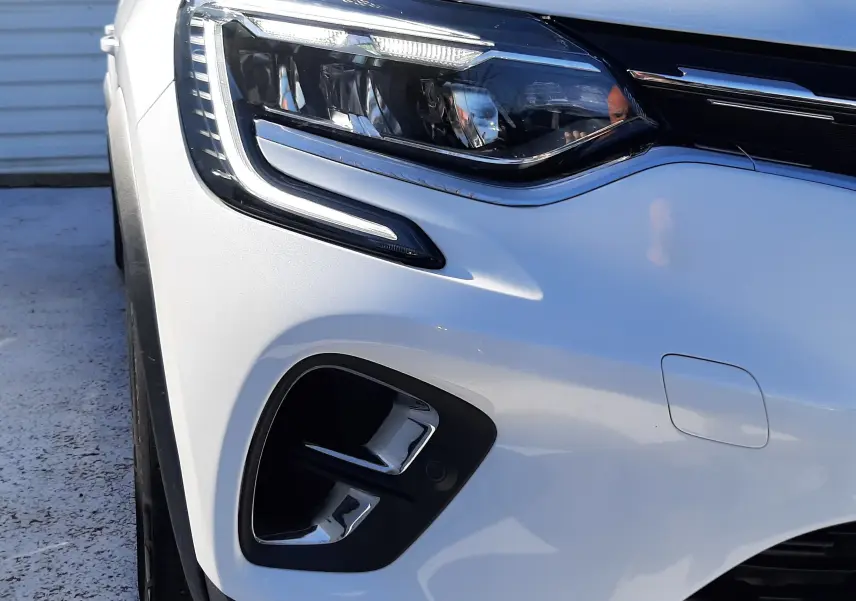 Gros plan sur l'avant gauche du Renault Captur E-Tech blanc nacré avec toit noir, mettant en valeur le phare LED et la calandre.