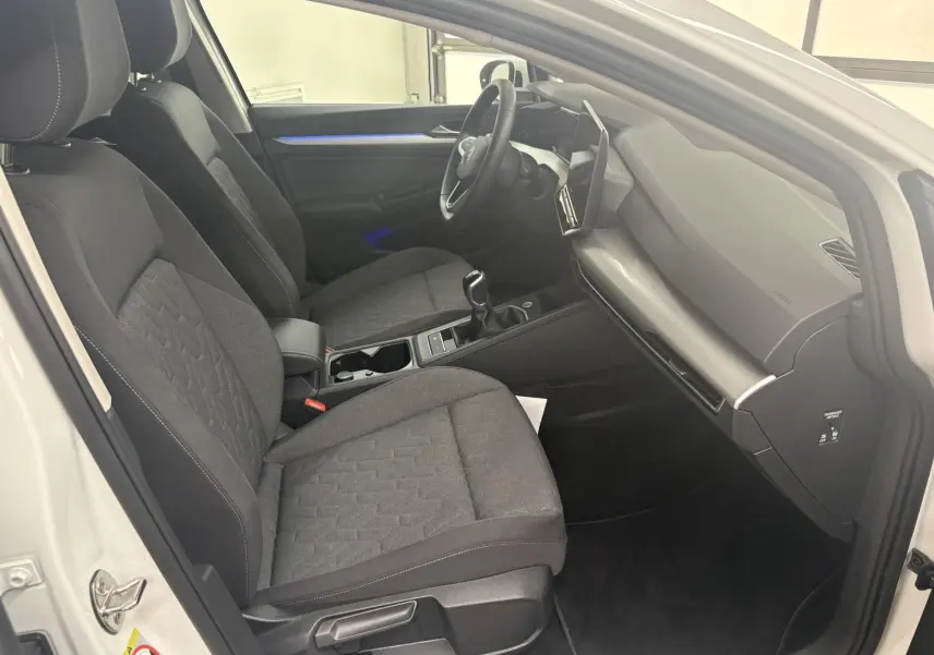 Vue intérieure côté conducteur de la Volkswagen Golf 1.5 eTSI blanche, montrant les sièges tissu gris et la console centrale avec boîte manuelle.
