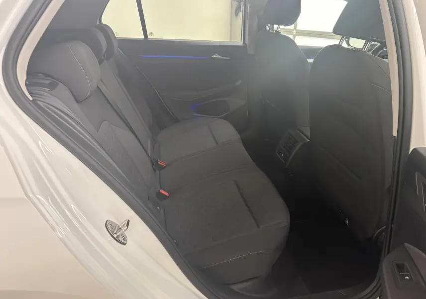 Vue intérieure arrière côté gauche d'une Volkswagen Golf 1.5 eTSI 2025 en blanc Oryx nacré, sièges tissu noirs et éclairage d'ambiance bleu.