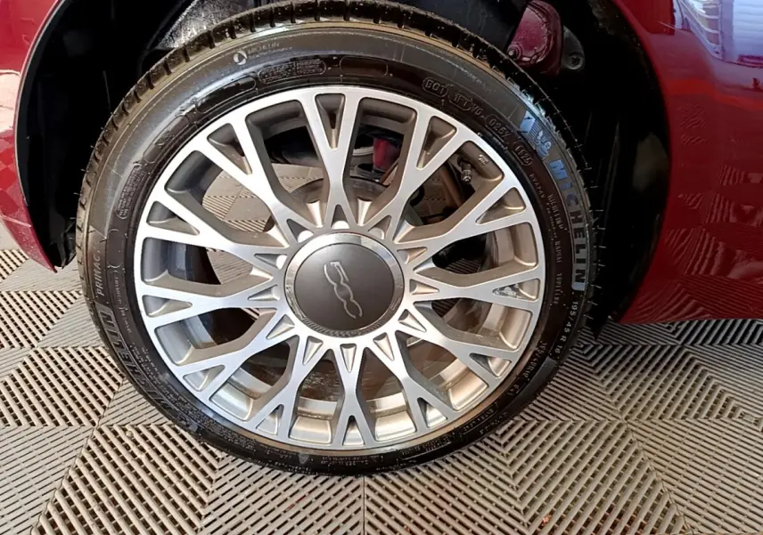 Gros plan sur la roue avant droite d'une FIAT 500 rouge, mettant en valeur la jante alliage argentée au design complexe.