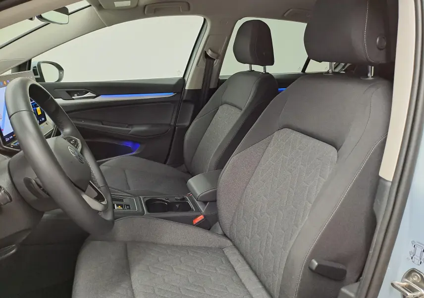 Vue intérieure côté conducteur de la Volkswagen Golf 1.5 eTSI 2025, sièges tissu gris avec éclairage d'ambiance bleu.