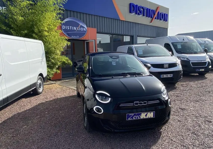 Vue avant d'une Fiat 500 électrique noire métal stationnée devant un concessionnaire avec d'autres véhicules blancs.