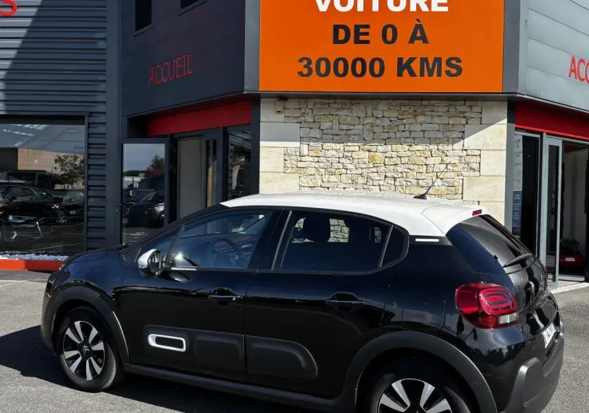 Vue 3/4 arrière droite d'une Citroën C3 noire à toit blanc avec protections latérales blanches, stationnée devant un bâtiment.
