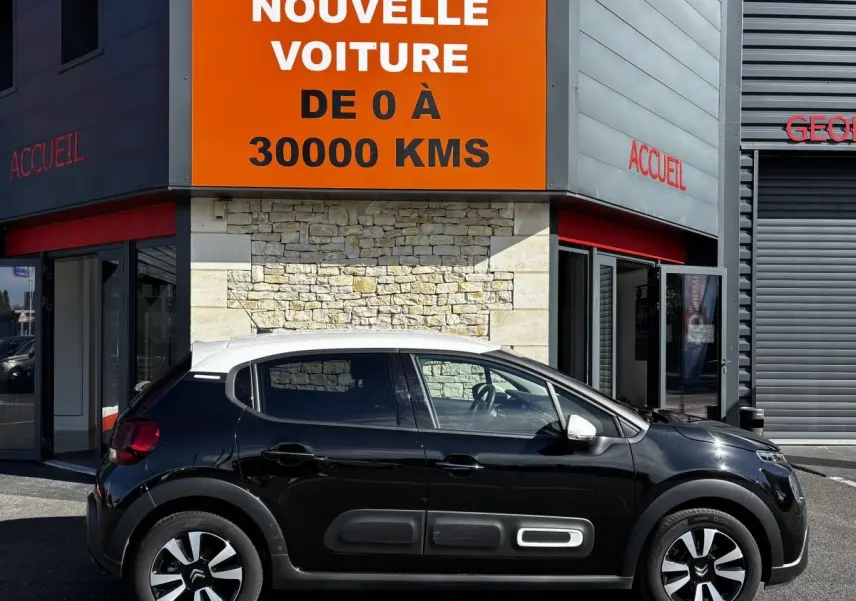Profil droit d'une Citroën C3 noire avec toit blanc, garée devant un bâtiment avec panneau orange "Votre nouvelle voiture".