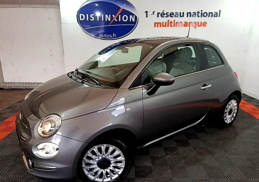 Fiat 500 Hybrid gris foncé vue 3/4 avant droit dans un showroom avec jantes argentées et phares allumés.