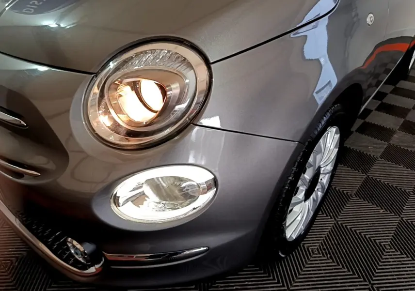 Gros plan sur l'avant droit gris foncé d'une Fiat 500 Hybrid avec phares allumés et jante alu visible.