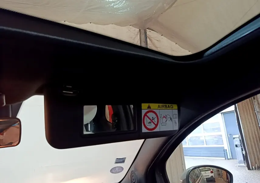 Vue intérieure sur le pare-soleil avec miroir et avertissement airbag, fenêtre ouverte côté droit, Fiat 500 gris foncé visible en reflet.