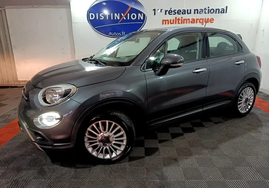 Fiat 500X gris foncé vue 3/4 avant droit, avec phares allumés et jantes alliage multibranches.