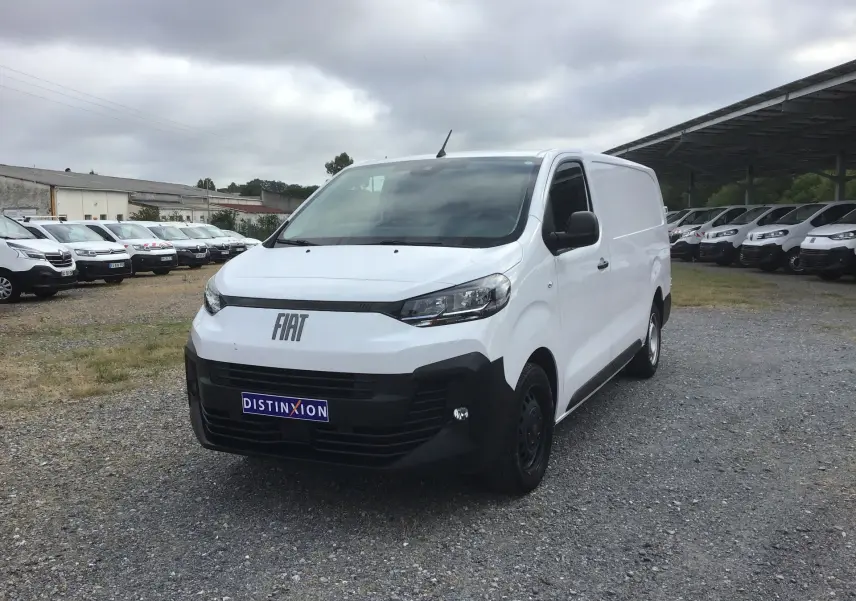 Vue 3/4 avant d’un FIAT Scudo Fourgon blanc 2024 avec pare-chocs noir et plaque de protection, stationné sur un sol gravillonné.