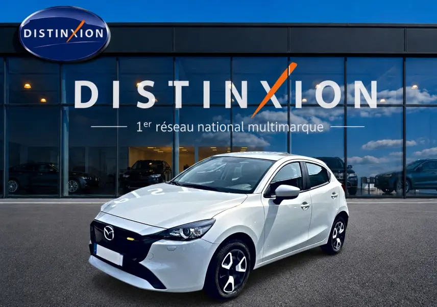 Vue 3/4 avant droite d'une Mazda2 blanche 2024 avec jantes noires et calandre noire devant un showroom Distinxion.