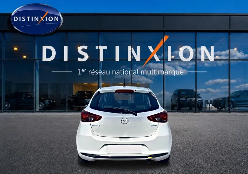 Vue arrière d'une Mazda2 blanche 2024 garée devant une vitrine avec logo Distinxion en arrière-plan.