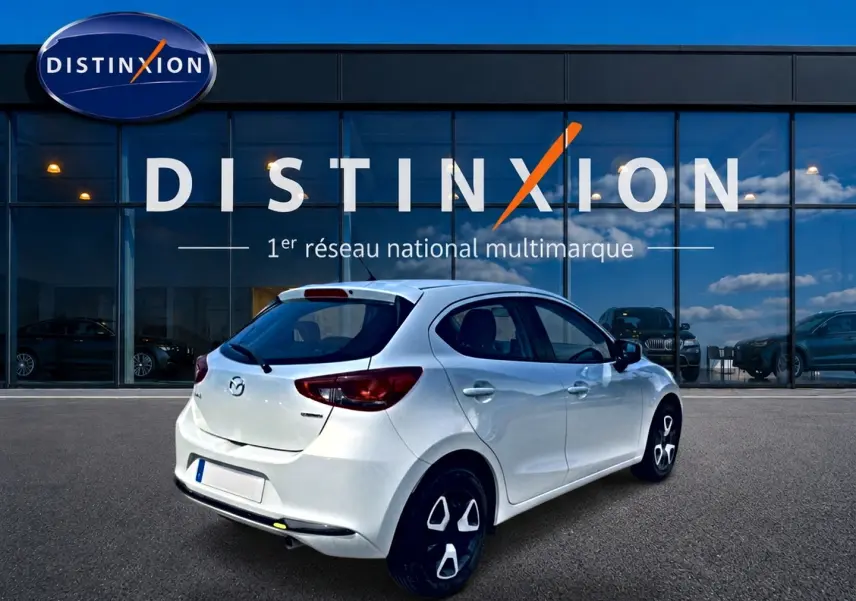 Vue 3/4 arrière droite d'une Mazda2 blanche 2024 garée devant une vitrine avec logo Distinxion.