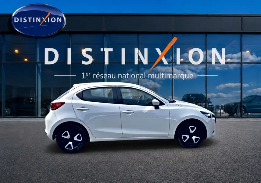 Vue latérale droite d'une Mazda2 blanche 2024 garée devant une vitrine avec logo Distinxion.