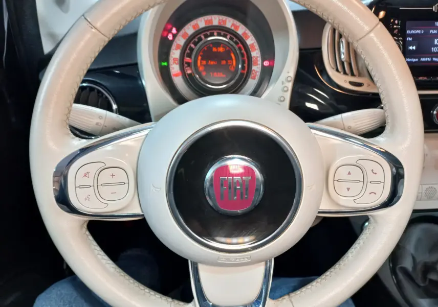 Vue rapprochée du volant beige de la Fiat 500 noire, avec commandes intégrées et tableau de bord lumineux en arrière-plan.