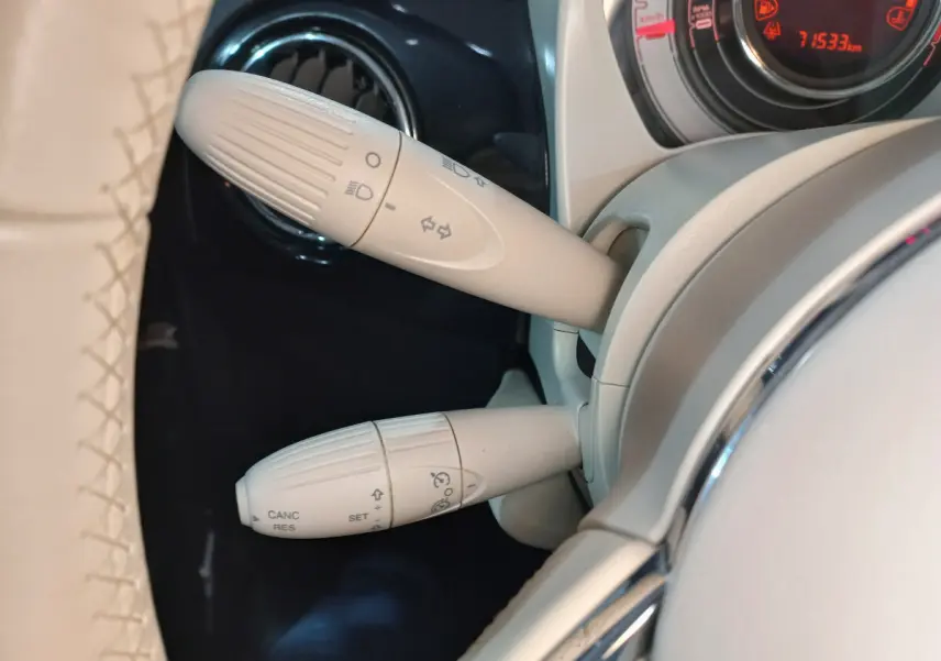 Gros plan sur les commandes beiges du volant cuir de la Fiat 500 Lounge noire, avec tableau de bord digital rouge en arrière-plan.