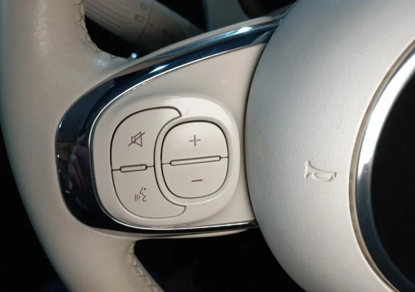 Gros plan sur les commandes de volume et de reconnaissance vocale sur le volant beige d'une Fiat 500 Lounge noire.