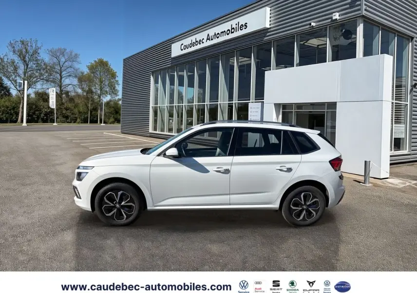 Profil côté gauche d'un Skoda Kamiq blanc 2025 garé devant un bâtiment moderne de concession automobile.