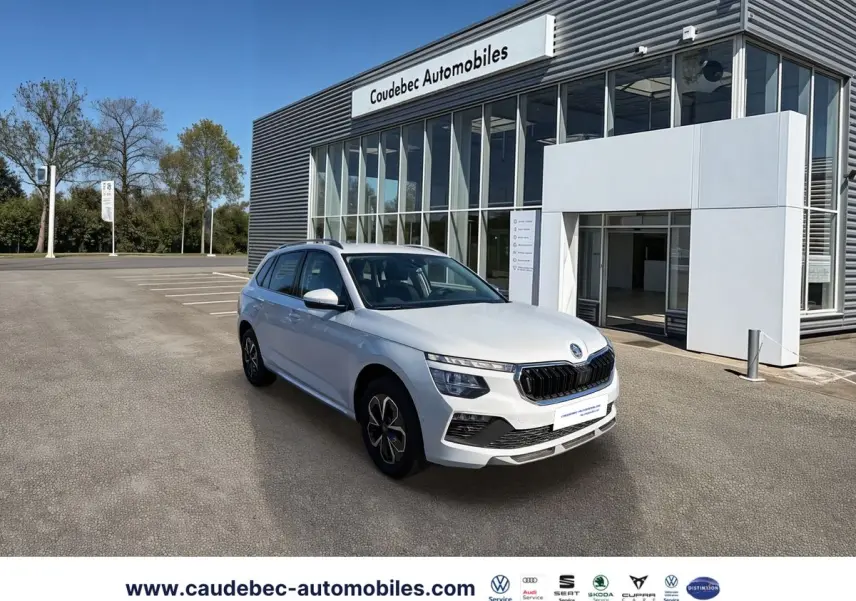 Vue 3/4 avant d'un Skoda Kamiq blanc stationné devant un bâtiment moderne de concession automobile.
