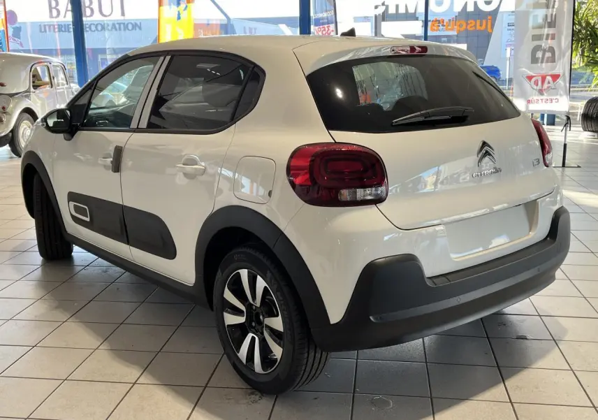 Citroën C3 blanc vue 3/4 arrière droit en showroom, avec protections noires et jantes alliage bicolores.