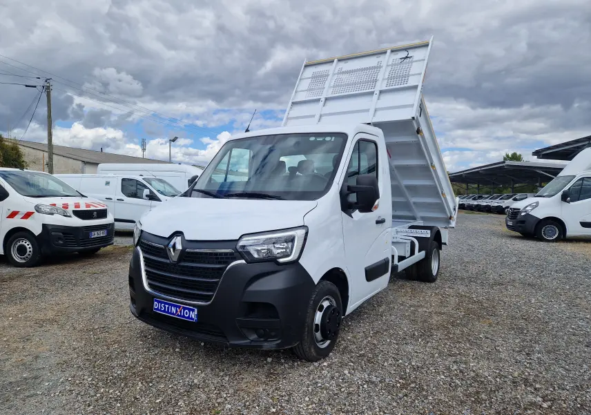 Renault Master Benne blanc vu en 3/4 avant droit, benne relevée et roues jumelées visibles sur terrain gravillonné.