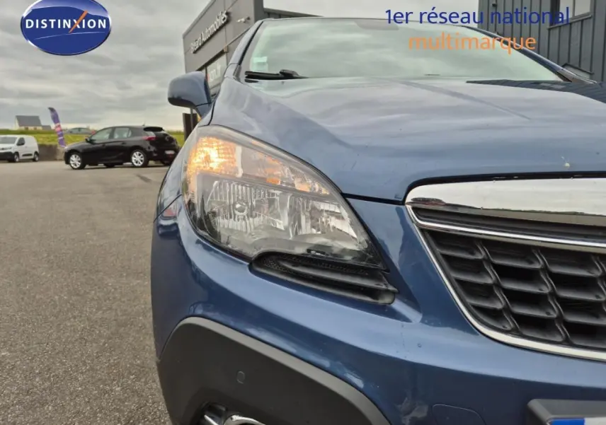 Gros plan sur l'avant gauche bleu outremer de l'Opel Mokka 2015 avec phare allumé et calandre chromée.