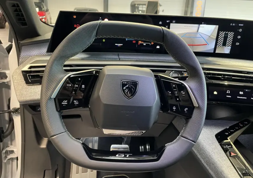 Vue rapprochée du volant cuir perforé avec badge GT du Peugeot 3008 Hybrid blanc, tableau de bord numérique en arrière-plan.