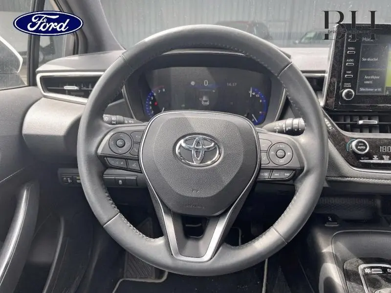 Vue intérieure centrée sur le volant cuir multifonction de la Toyota Corolla blanche, avec tableau de bord digital et écran tactile.