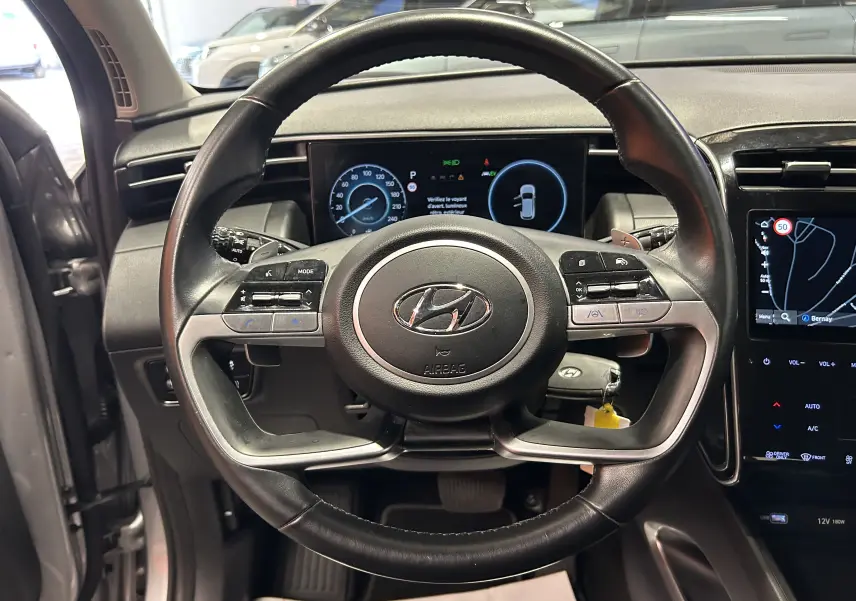 Vue intérieure centrée sur le volant cuir noir du Hyundai Tucson Hybrid 2022 avec tableau de bord numérique et écran GPS.
