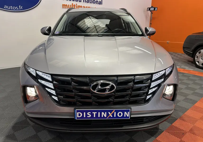 Vue avant d'un Hyundai Tucson hybride gris Shimmering Metal avec calandre noire et feux LED allumés en intérieur showroom.