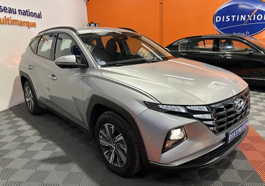 Vue 3/4 avant d'un Hyundai Tucson hybride gris Shimmering Metal avec calandre distinctive et feux allumés en intérieur showroom.