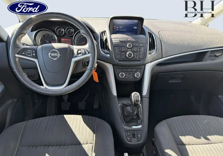 Vue intérieure avant de l’Opel Zafira Tourer 2013, tableau de bord et volant avec logo Opel, sellerie tissu gris.
