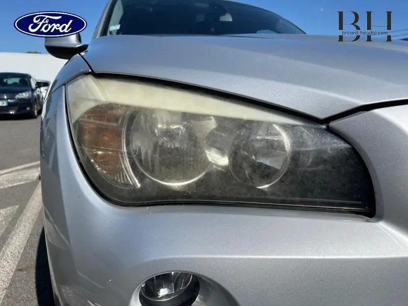 Gros plan sur le phare avant gauche d’un BMW X1 gris métallisé, avec le pare-chocs et le feu antibrouillard visibles.