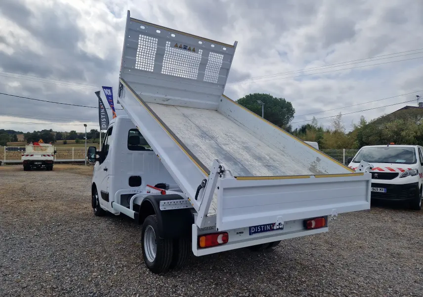 Vue 3/4 arrière droite d’un Renault Master blanc avec benne basculante relevée et roues jumelées sur terrain extérieur.